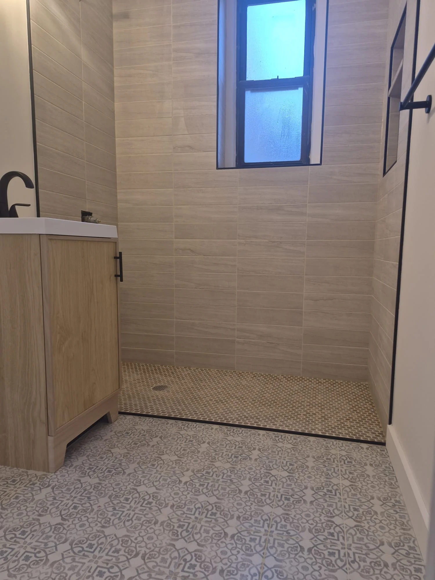 Pre-war master bath detail — wood-grain porcelain wall tile, hex pebble shower floor, light wood vanity